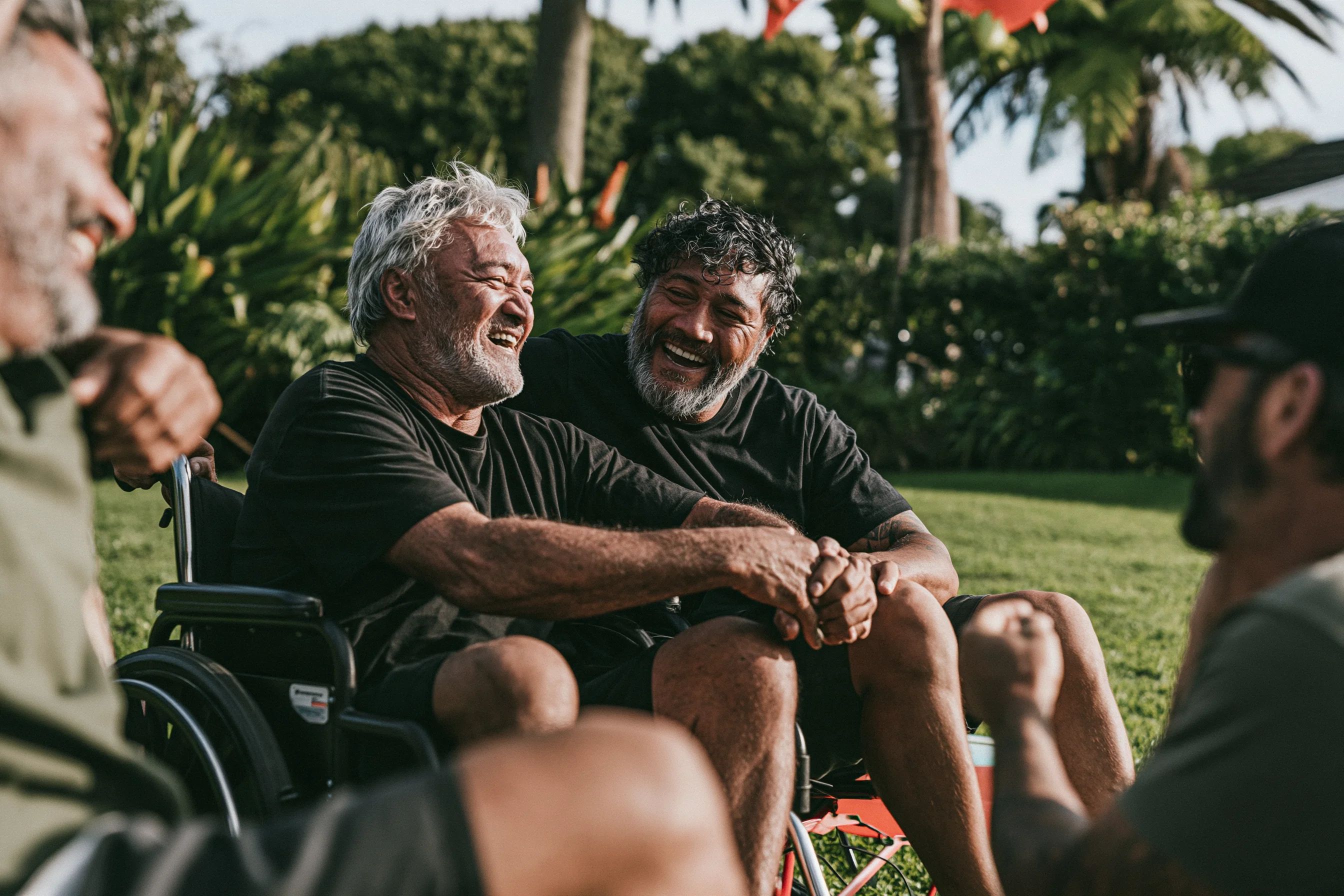 An older man in a wheelchair laughing with friends in a sunny backyard, hands clasped