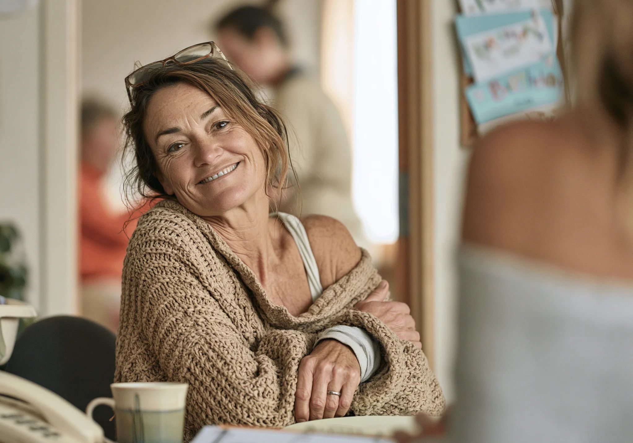 A rest-home owner in her sixties smiling across her desk, glasses pushed up, a mug and phone beside her